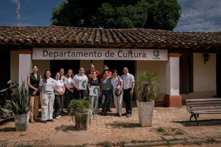 Treinta personas sonríen en exterior, frente al edificio del Departamento de Cultura, vistiendo ropa casual en un ambiente festivo.