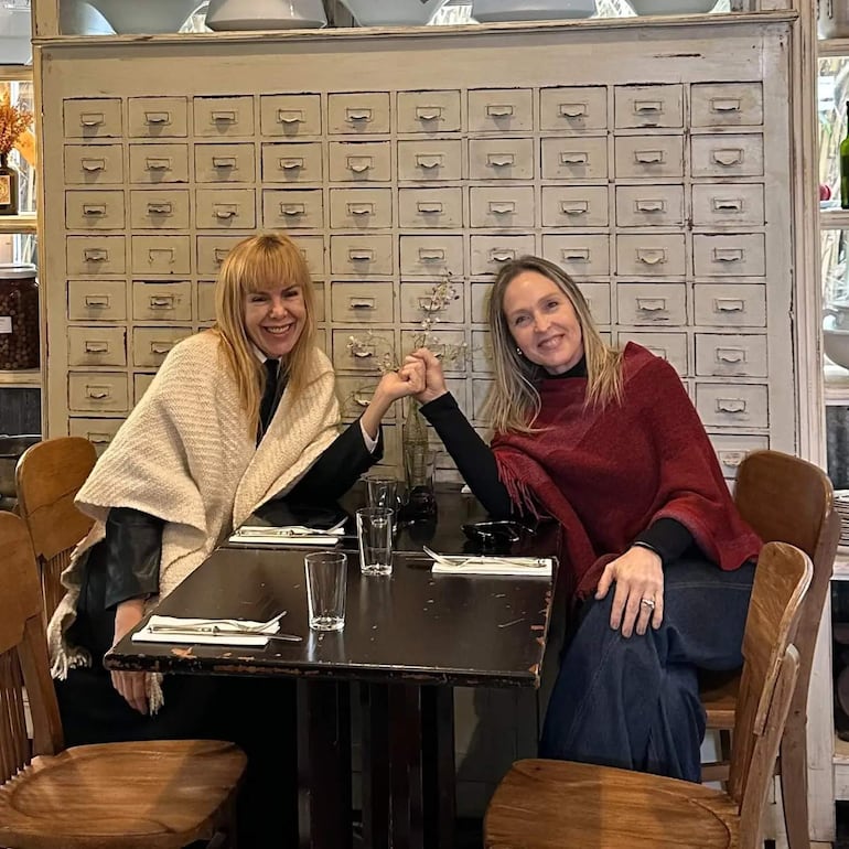 Dos mujeres sonrientes entrelazan sus manos en una mesa de café, una lleva chal rojo y la otra un abrigo claro, en un ambiente acogedor.