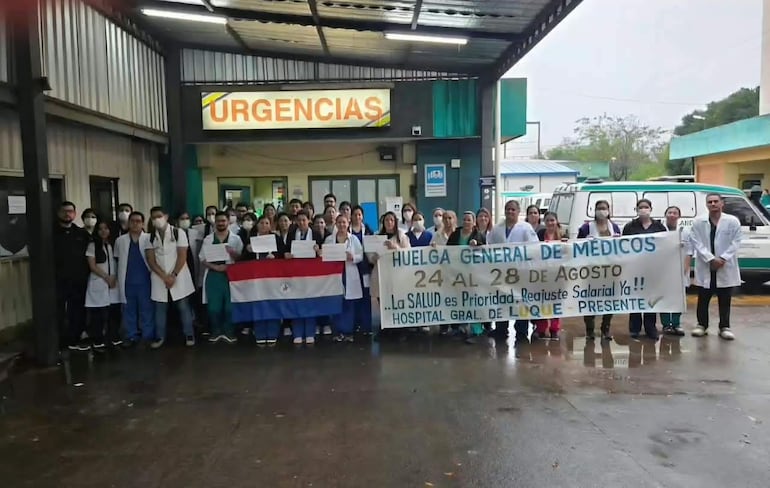 Grupo de médicos en batas y camisetas coloridas sosteniendo pancarta de huelga frente al hospital. Atmosfera tensa pero esperanzadora.
