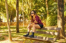 Luce con cabello rizado, camiseta burdeos y pantalones cortos, sentada en un banco rodeada de árboles.