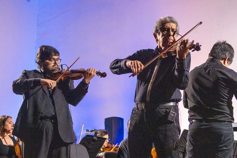 Músicos tocando violines en el escenario. Uno con saco negro y gafas, otro con cabello canoso y camisa negra, bajo luces azules y blancas.