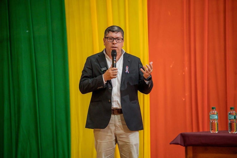Daniel Pereira Mujica en traje oscuro gesticulando al micrófono, rodeado de cortinas en colores verde, amarillo y naranja.