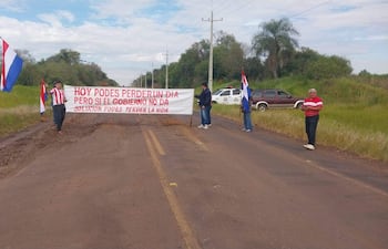 Dos personas con banderas paraguayas y un hombre con camiseta a rayas sostienen un cartel de protesta en la ruta rodeada de vegetación.