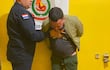 Hombre con chaqueta verde aparentemente sometido por un oficial de policía en un ambiente de detención con pared amarilla.