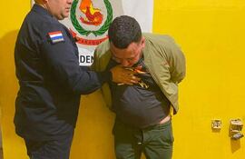 Hombre con chaqueta verde aparentemente sometido por un oficial de policía en un ambiente de detención con pared amarilla.