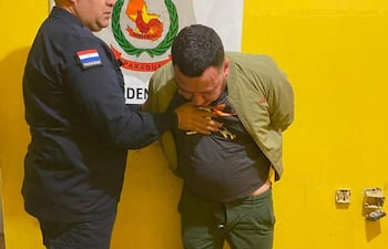 Hombre con chaqueta verde aparentemente sometido por un oficial de policía en un ambiente de detención con pared amarilla.