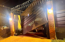 Portón metálico caído y cerca de alambre de púas en un ambiente oscuro, iluminado por faroles, sin personas visibles.