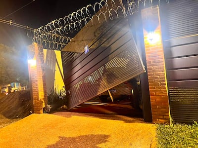 Portón metálico caído y cerca de alambre de púas en un ambiente oscuro, iluminado por faroles, sin personas visibles.