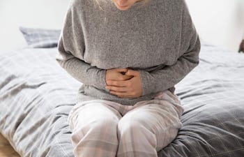 Persona en suéter gris y pijama de cuadros en la cama, sosteniéndose el abdomen con expresión de malestar en un dormitorio.