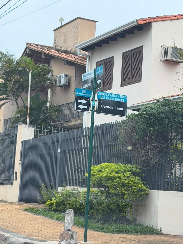 Calle en Asunción con casas y carteles que instan a usar casco en moto. Cielo nublado y jardín visible.