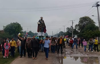 Multitud en procesión con banderas, figuras religiosas y clima nublado en Puerto Sastre.