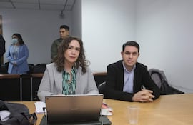 Hernán Rivas, hombre de cabello corto y camisa a cuadros, conversa con mujer de blusa de rayas verdes en sala formal.