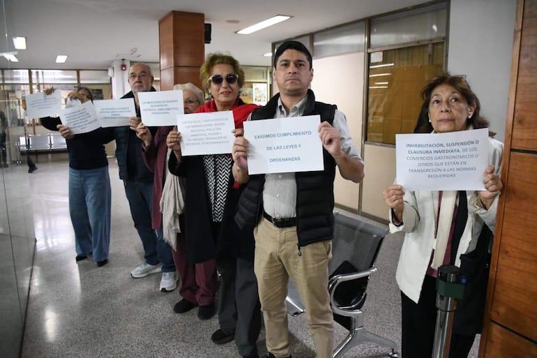 Seis personas de pie en un ambiente formal sosteniendo carteles, algunas visten abrigo, y una lleva gafas de sol.