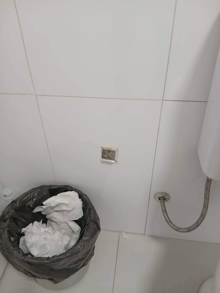 Vista de un baño con cesto de basura negro lleno de papel higiénico arrugado y un inodoro visible.