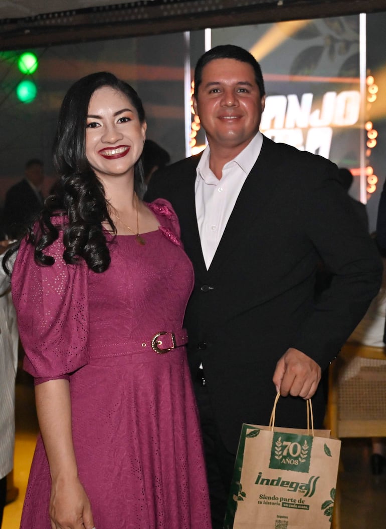 Mujer con vestido morado y cabello rizado sonríe junto a hombre en camisa blanca y saco negro en ambiente festivo.