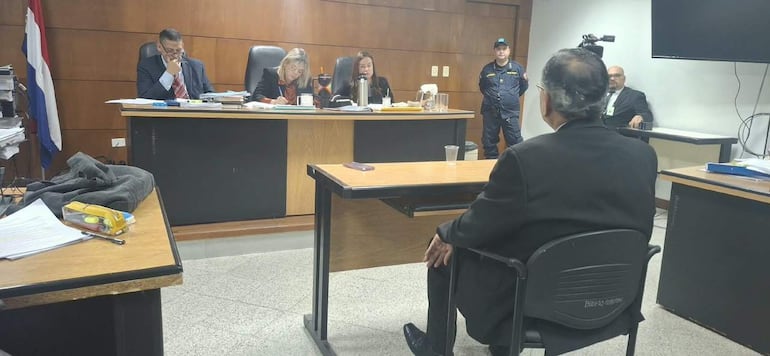 Óscar Rodríguez Kennedy, de espaldas, escucha atento en audiencia judicial, rodeado de personas en un ambiente formal y documentos sobre la mesa.