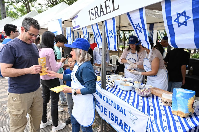 Mujer con gorro azul entrega folleto a hombre de camisa azul, rodeados de jóvenes y una carpa con decoraciones israelíes.
