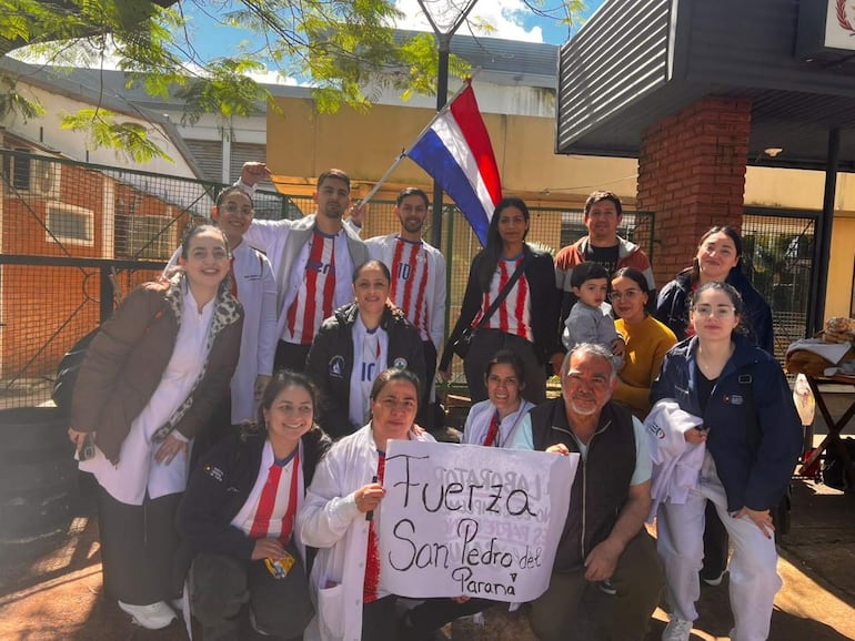 Grupo de bioquímicos sosteniendo un cartel de apoyo, con camisetas de franjas rojas y blancas, en un día soleado frente a un hospital.