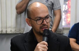 Hombre calvo y con gafas habla al micrófono, vistiendo abrigo negro, rodeado de personas en un ambiente formal.