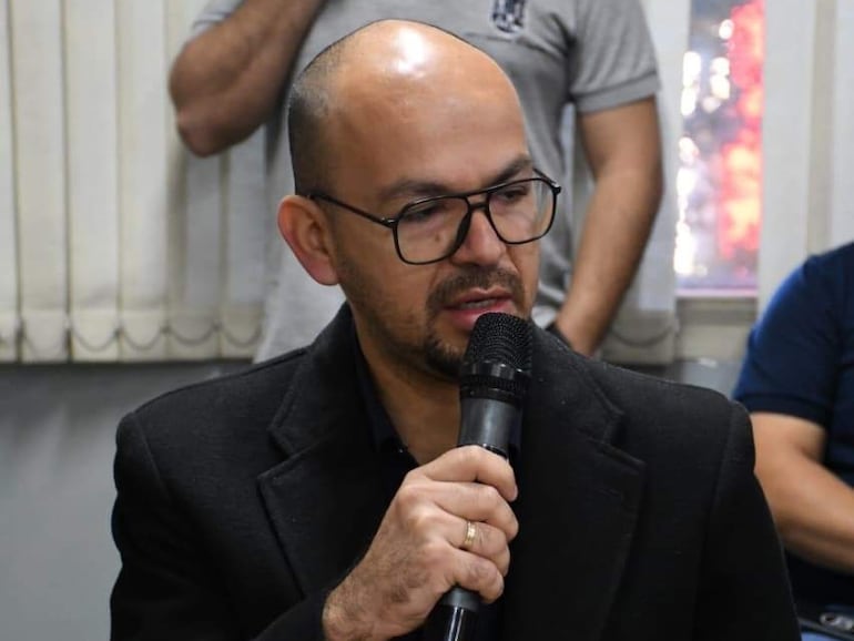Hombre calvo y con gafas habla al micrófono, vistiendo abrigo negro, rodeado de personas en un ambiente formal.
