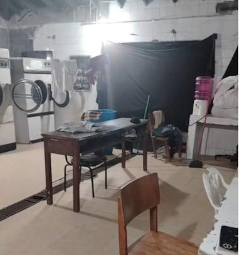 Interior desordenado de lavandería sin personas. Máquinas de lavar y secar a la izquierda, sillas y mesa de madera en el centro.