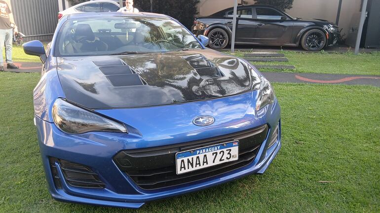 Automóvil deportivo azul con capó negro estacionado en césped, sin personas visibles en la imagen.