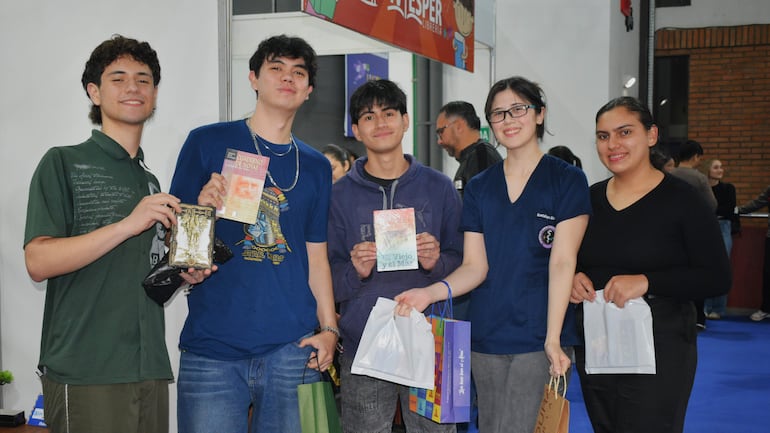 Cinco jóvenes sonríen en evento literario, uno sostiene un cuaderno azul y otro un libro, en un ambiente con pared de ladrillos.