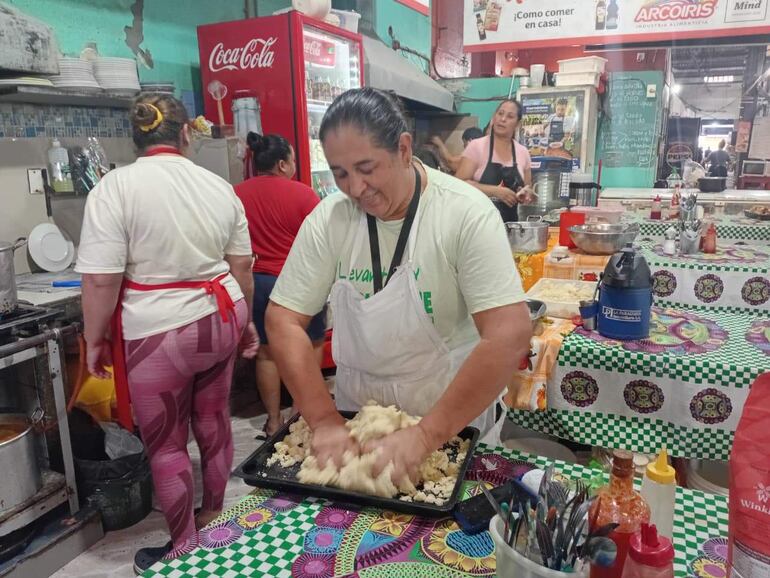 Cuatro mujeres en cocina industrial, una en delantal blanco amasando, otras colaborando con equipamiento visible y ambiente activo.