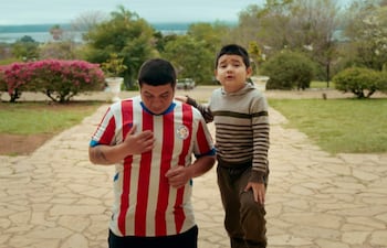 Claudio Rejala con camiseta roja y blanca, junto a su hijo en suéter de rayas, en un parque con vegetación colorida.