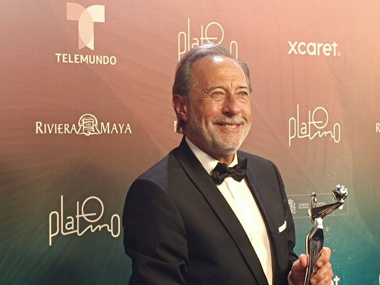 Guillermo Francella, con traje negro y pajarita, sonríe mientras sostiene un trofeo, rodeado de logotipos de Telemundo y el evento Platino.