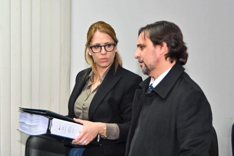 Melissa Ramírez con blazer negro y blusa gris sostiene un expediente, mientras Jorge Sabaté escucha en abrigo negro y camisa blanca.