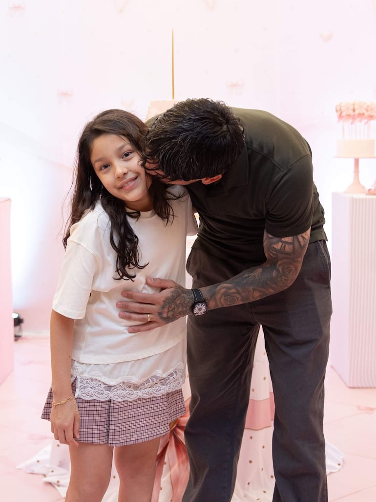 Hombre tatuado con camiseta oscura sonríe a una niña con blusa blanca y falda de cuadros, en un fondo decorativo rosa.