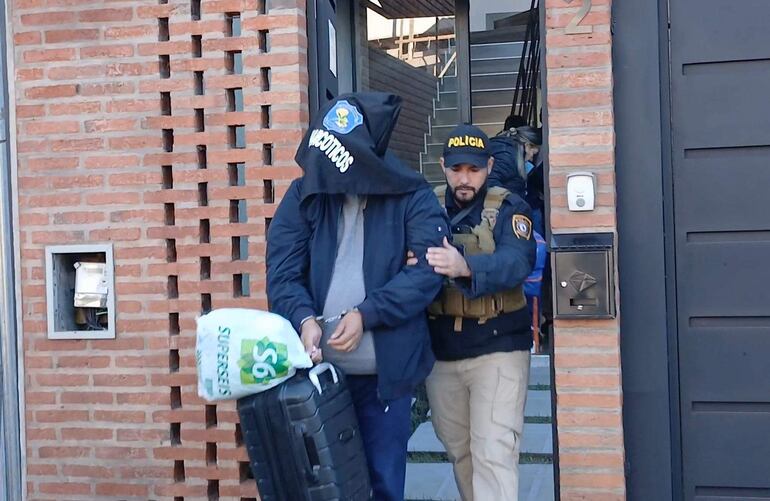 Hombre con capucha negra siendo detenido por agente policial en un operativo, sosteniendo un maletín y una bolsa plástica.