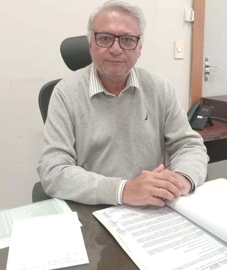 Hombre con gafas y suéter gris claro revisa hojas en su oficina, rodeado de documentos y un teléfono de escritorio.