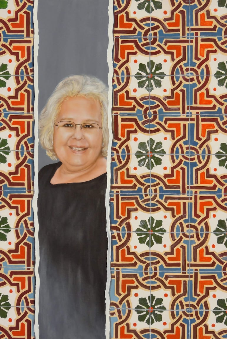 Mujer con cabello rizado y canoso, gafas y camisa negra sonríe frente a un fondo decorado en colores vivos.