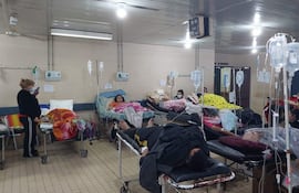 Mujer de pie a la izquierda, tres pacientes en camas visibles, ambiente hospitalario con paredes beige y luz artificial.
