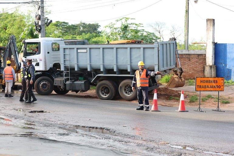 Cuatro hombres trabajadores en chalecos reflectantes y cascos amarillos, uno señala mientras un camión y excavadora están de fondo.