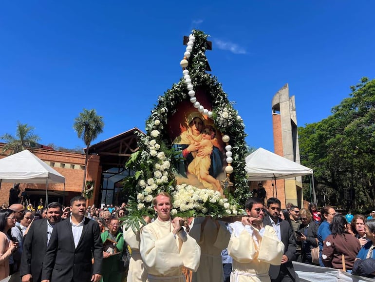 Hombres vestidos de blanco sosteniendo una imagen de la Virgen María entre una multitud formal y colorida bajo un cielo soleado.