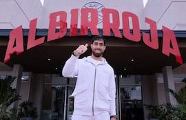 La imagen muestra a una persona de pie frente a un edificio que tiene el letrero "ALBIR ROJA" claramente visible. La persona, que se presenta con una vestimenta deportiva de color gris, sostiene un gesto con el pulgar hacia arriba, sugiriendo satisfacción o aprobación.
No hay otras personas visibles en la escena. La ambientación es exterior, frente a un complejo que parece relacionado con el fútbol, dado el nombre y la simbología asociada con la selección nacional de Paraguay.
El entorno incluye plantas decorativas y un acceso al interior del edificio. Dado el contexto, se puede inferir que la persona podría ser un futbolista o figura pública vinculada al deporte local, aunque no se puede identificar con alta confianza. No hay texto adicional visible en la imagen.