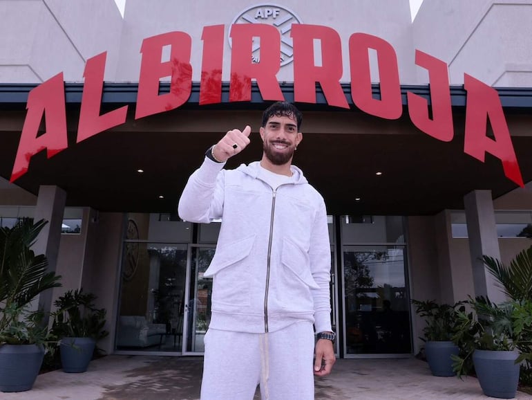 La imagen muestra a una persona de pie frente a un edificio que tiene el letrero "ALBIR ROJA" claramente visible. La persona, que se presenta con una vestimenta deportiva de color gris, sostiene un gesto con el pulgar hacia arriba, sugiriendo satisfacción o aprobación.
No hay otras personas visibles en la escena. La ambientación es exterior, frente a un complejo que parece relacionado con el fútbol, dado el nombre y la simbología asociada con la selección nacional de Paraguay.
El entorno incluye plantas decorativas y un acceso al interior del edificio. Dado el contexto, se puede inferir que la persona podría ser un futbolista o figura pública vinculada al deporte local, aunque no se puede identificar con alta confianza. No hay texto adicional visible en la imagen.