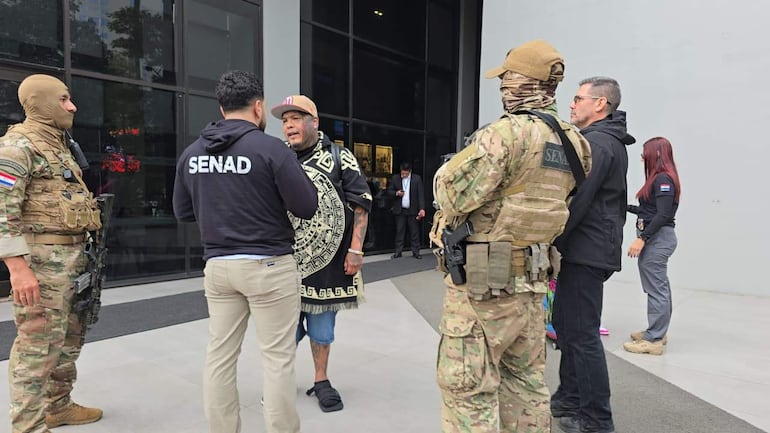 Hombres de seguridad con uniformes militares cerca de Troy Vasquez, quien viste sudadera negra, conversando con otro individuo en un entorno urbano.