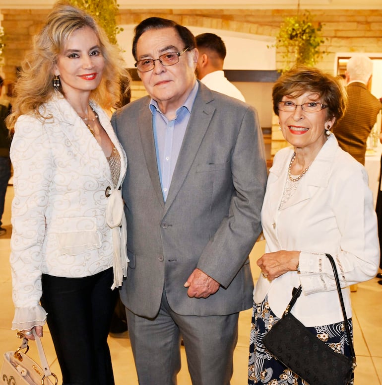 Mujer rubia sonriente con bolso, hombre de traje gris con gafas y mujer mayor de atuendo blanco, todos en evento social.