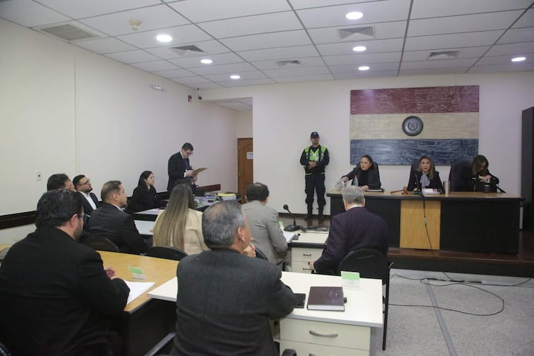 Un grupo de personas en sala judicial, un hombre de pie lee documentos frente a tres juezas atentas.