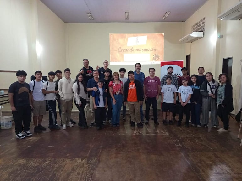 Grupo sonriente de jóvenes y niños en un salón, con camisetas variadas, participando en la creación de canciones frente a una proyección.