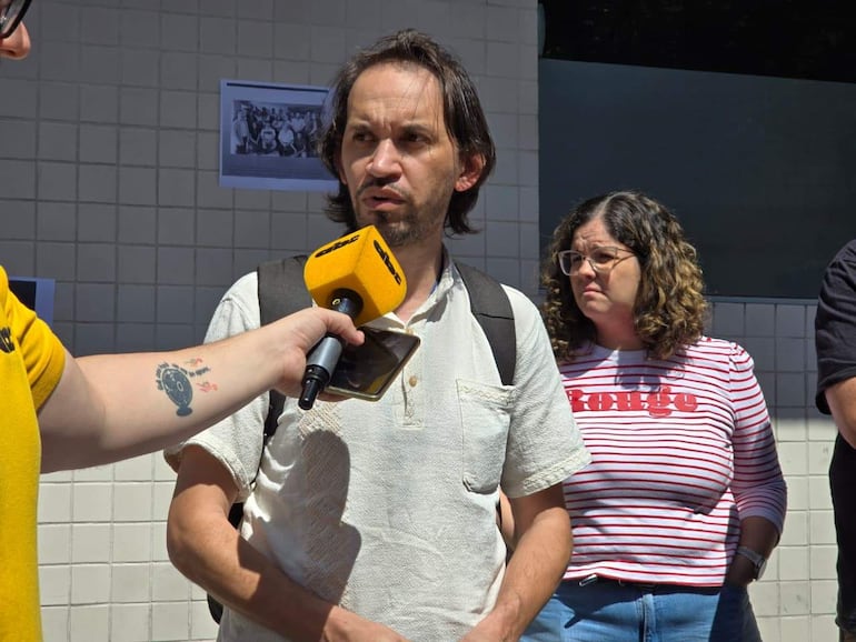 Persona con micrófono y camiseta amarilla, orador con camisa blanca y mujer de camiseta a rayas observa atentamente en un evento al aire libre.