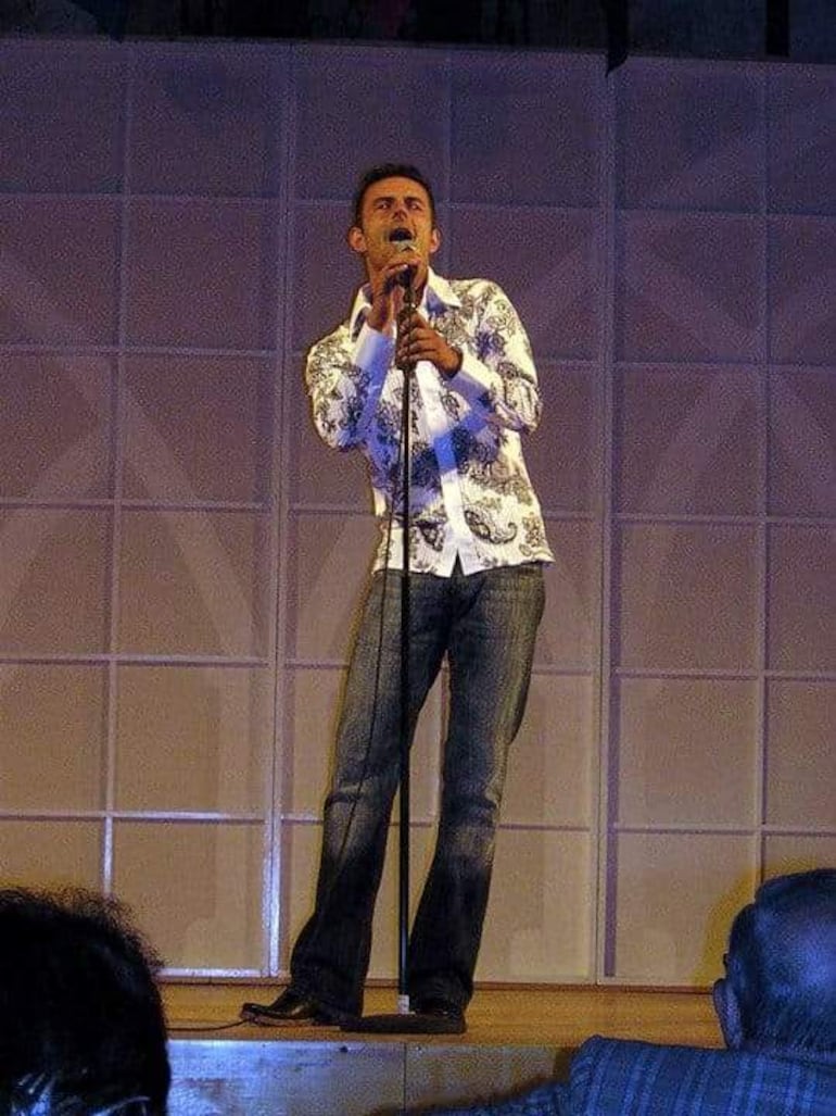 Hombre de pie con micrófono, vestido con camisa blanca con patrones oscuros y jeans, canta en un escenario frente a una audiencia.