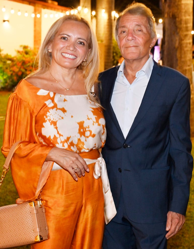 Mujer rubia sonriente con vestido naranja y hombre canoso en traje oscuro, rodeados de luces decorativas y vegetación.
