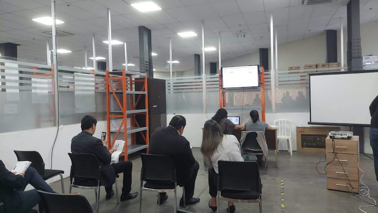 Seis personas, incluyendo una mujer de espaldas, en sala sobria con estanterías anaranjadas y pantallas mostrando información.