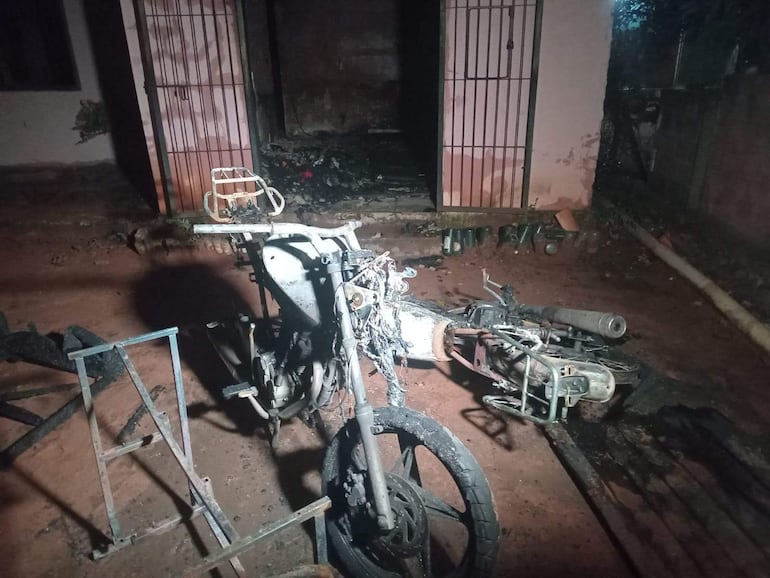Motocicleta quemada y caída en terreno de tierra, con estructura de edificación sucia de fondo.