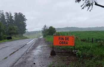 Carretera mojada en entorno rural con cartel naranja que dice 'FIN DE OBRA', rodeada de vegetación y colinas suaves.